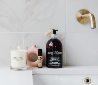 A white jar glass candle which is lit, sitting on the edge of a sink in a bathroom with various bathroom items sitting next to it. 