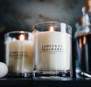 A white candle in a glass jar without a lid with the James and Co Fragrance logo on the front. It is lit, against a dark background.