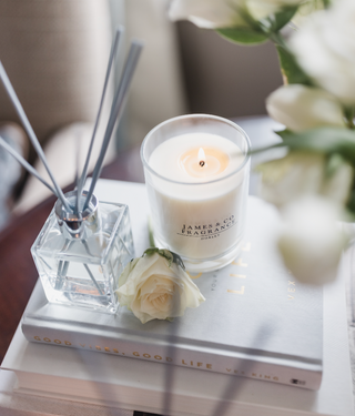 A white glass jar candle which is lit, sitting next to glass reed diffuser with grey reeds on top of a pile of books.