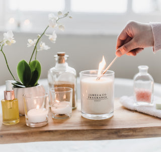 A white glass jar candle being lit by a match. In the background is a bathroom with various objects. 