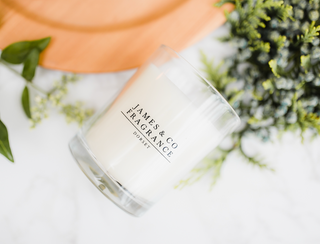 White glass candle laid on its site with James and Co Fragrance logo on the front. In the background is various foliage.