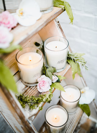 Four clear glass candles in open top jars, sitting on a shelving unit with flowers scattered surround. 