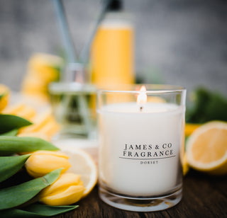 A lit glass jar candle sitting on a wooden board with various flowers and tulips scattered around.