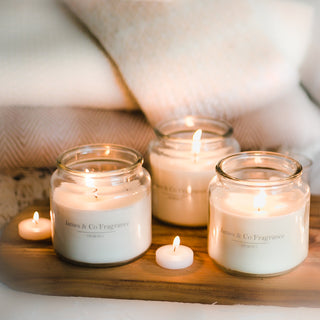 Three lit white jar candles sitting on  a wooden surface with tealights scattered around.