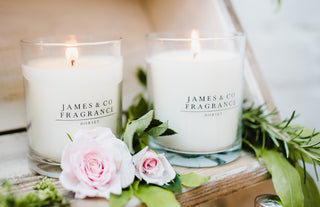 Two lit glass jar candles, sitting on a wooden shelf with flowers scattered around them.