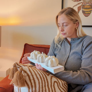 Woman sitting on a couch holding a tray with small pumpkins, surrounded by warm lighting.
