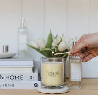 A white female hand, holding a match, lighting a white glass jar candle. 