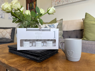 A trio of NO.1 white fragranced reed diffuser set sitting on slate place mats. Directly behind is a fresh bunch of roses. and to the right is a mug of a hot cup of tea. 