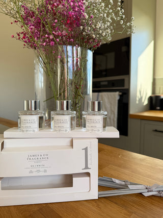 Sitting on a kitchen island is a trio of reed diffusers, outside of the white packaging displaying the fragrance. The scent is NO.1 white. Behind is a vase of fresh Gypsophila (Baby's-breathe) with colours of deep pink, and white. 