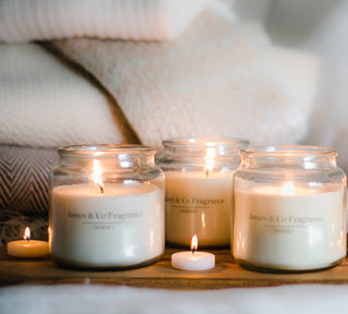 A range of luxury candles are lit, places on a wooden table beside a blanketed sofa.