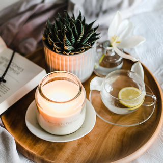 A lit glass jar candle sitting on a wooden board with a cup of tea, a plant and a reed diffuser.