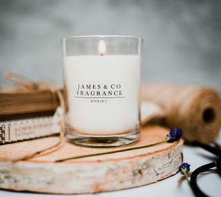 A lit glass jar candle with the J&Co logo on the front sitting on a wooden board.