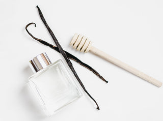 Glass bottle laying on a white background next to vanilla pods and honey stick