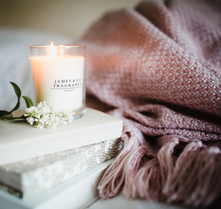 Candle labeled 'James & Co Fragrances' on a surface with a pink blanket and flowers in the background