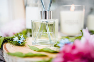Glass diffuser bottle labeled 'James & Co' with reeds on a wooden surface with flowers and candles in the background.