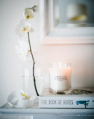 Candle with 'James  & Co Fragrance' on a surface with white orchids and a book.