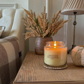 A lit candle, sat on a wooden table, with autumnal flowers in the background and a lamp.