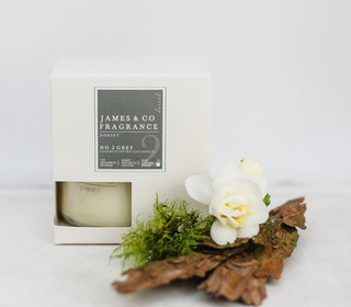 A candle in a white box with a grey label that reads 'No.2 Grey'. In front of the candle is a piece of bark, and a white flower. 