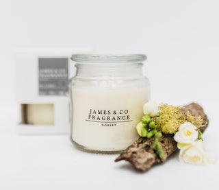 A large glass jar candle with a lid, sitting next to its box and a piece of bark. 