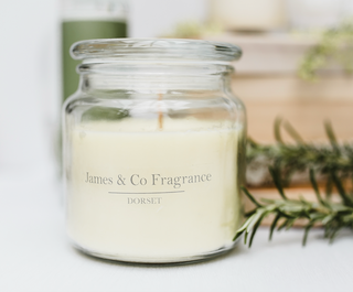 A glass jar white candle with a lid sitting on a white surface with a bottle of refill oil in the background. 