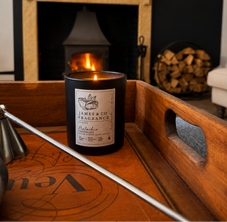 A black scented candle with a white label reading James & Co Fragrance sits on a wooden tray in a cozy room with a lit fireplace and wood stack in the background.