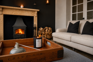 A cozy living room scene featuring a lit black metal fireplace insert with a wood fire, a cream sofa with black pillows, a stack of firewood, and a wooden tray in the foreground holding a candle.