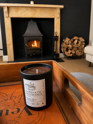A black scented candle with a white label reading James & Co Fragrance is placed on a wooden tray in a cozy living room setting, with a lit wood-burning fireplace in the background.