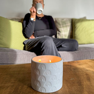 A ceramic candle in white, the ceramic is patterned with rainbows all over. The candle is lit and sitting on a wooden table. Behind a lady is enjoying a cup of tea.