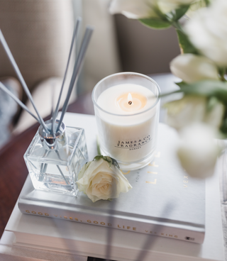 A reed diffuser and candle both in clear glass jars, the candle is lit. There are white rose surrounding the image. The objects are displayed on a book on a coffee table.