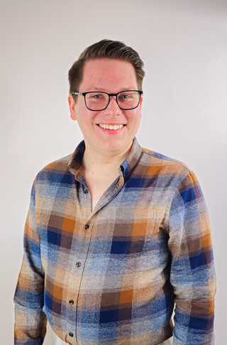 A young male, he has black framed glasses with a big smile. He has brown hair. The man is wearing a checked shirt in the rustic colours. 